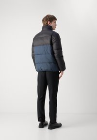 Puffjakke designet med svarte og marineblå fargeblokker, høy krage, glanset finish og horisontalt quiltet tekstur; kombinert med svarte bukser.