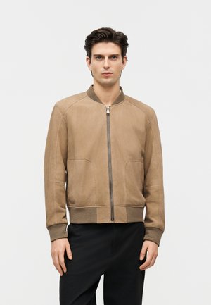 Mees seisab otse vaataja suunas, kandmas helepruuni bomberjopet, millel on tõmblukk ja soonikkoes varrukasääred, ning musti pükse, valgel ühevärvilisel taustal.