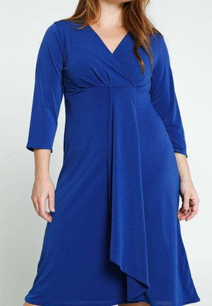 Vrouw draagt een koningsblauwe jurk tot op de knie met driekwart mouwen en een V-hals wikkelbodice met geplooide details.