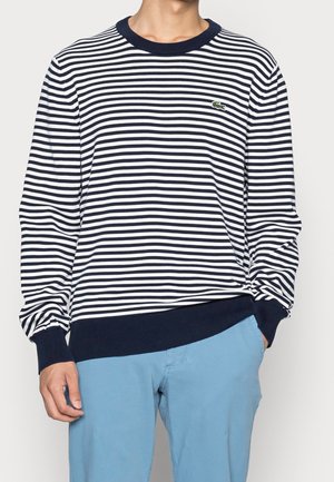 Man draagt een marineblauw-wit horizontaal gestreepte trui met lange mouwen, marineblauwe manchetten en zoom, gecombineerd met lichtblauwe broek.