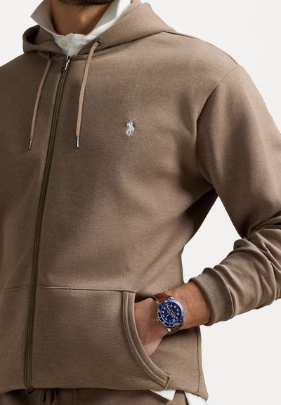 Brūns zip-up hoody ar baltu logo, ar auklītēm un sānu kabatām. Valkāts ar zilu pulksteni un brūnu ādas siksnu.