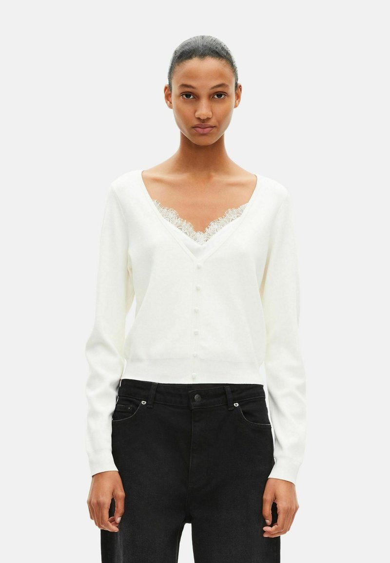 Hvid cardigan med dyb V-hals, dekoreret med blonder under halslinien og små knapper foran. Bruges sammen med sorte jeans.