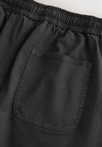 Pantalon avec taille élastique noire et une poche arrière carrée unique avec des coutures visibles sur un fond uni.