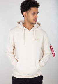 Haut en molleton beige clair fabriqué dans un tissu doux, doté d'une poche avant, de cordons de serrage et d'une poche zippée sur le côté avec une étiquette rouge.