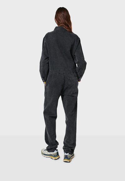 Schwarzer Denim-Overall mit langen Ärmeln, elastischem Bund und seitlichen Taschen; getragen mit grauen Laufschuhen mit gelben und schwarzen Akzenten.