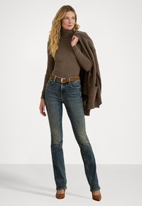 Camisola de gola alta castanha, jeans azuis descontraídos com um tom desbotado, cinto de couro castanho e sapatos de salto castanhos; cardigã de lã pousado sobre o ombro.