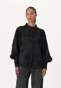 Camicia nera ricamata con maniche a palloncino, caratterizzata da motivi floreali e una chiusura a bottoni. Realizzata in un tessuto leggero e trasparente.
