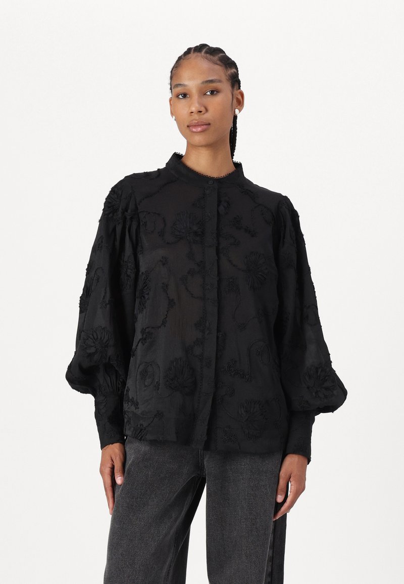 Camicia nera ricamata con maniche a palloncino, caratterizzata da motivi floreali e una chiusura a bottoni. Realizzata in un tessuto leggero e trasparente.
