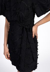 Schwarzes Kleid mit floraler Stickerei, Wickeldesign, kurzen Ärmeln und einer gebundenen Taillendetail. Der Stoff hat eine strukturierte Optik.