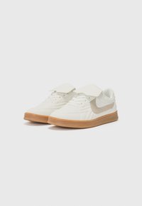 Baskets blanches avec une surface texturée, dotées d'une accent beige et d'une semelle en caoutchouc. Le design comprend une sangle en Velcro pour un ajustement sécurisé.