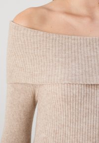 Beige off-shoulder ribbed sweater with a wide neckline and long sleeves. Soft knit texture with vertical stripes throughout.