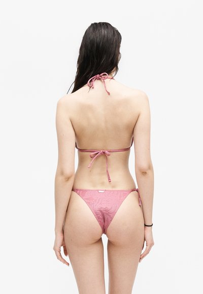 Γυναίκα που στέκεται με την πλάτη στην κάμερα, φορώντας ένα ανοιχτό ροζ υφασμάτινο μπικίνι με δεμένους ιμάντες σε λευκό φόντο.