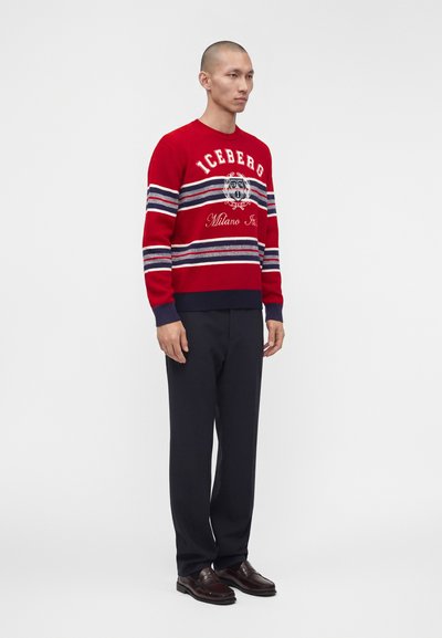 Homme debout portant un pull rouge à manches longues avec des rayures blanches et bleues et un pantalon noir, chaussures en cuir marron, sur fond blanc uni.