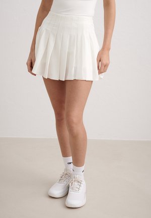 Kvinde iført et hvidt plisseret skørt, hvide træningsstrømper og hvide sneakers, stående mod en ensfarvet baggrund.