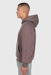 Profil osoby w brązowej bluzie z kapturem i jasnoniebieskich dżinsach z przetarciami, stojącej na tle jednolitego szarego tła.