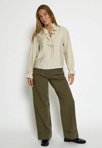 Beige ruffled blouse with a tie neck, textured fabric, and long sleeves, paired with olive-green wide-leg pants and black shoes.