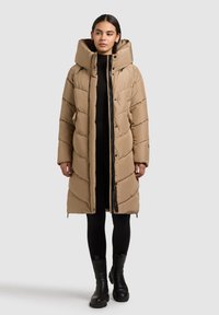 Béžový péřový kabát s kapucí, prošitý design, přední zip a boční kapsy. Nošený přes černý outfit a doplněný černými botami.