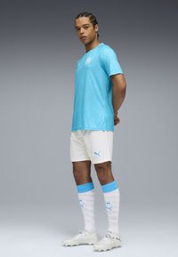 Gaiši zils sporta krekls ar maziem oranžiem punktiņiem, balti šorti, baltas zeķes ar zilām maliņām un sudraba futbola botas. Redzams PUMA logo.