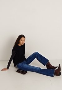 Musta neulepaita, vaaleansiniset suorat farkut, ruskeat nilkkurit. Malli istuu tumman laukunkl kanssa. Yksinkertainen tausta.