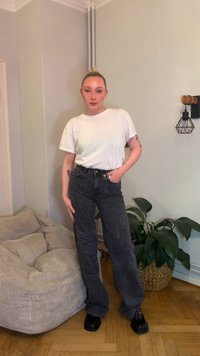 Bela majica s kratkimi rokavi v kombinaciji s širokimi kavbojkami z vzorcem črno-sivih zebri in črnimi škornji na platformo, stoji ob svetlo obarvani steni.