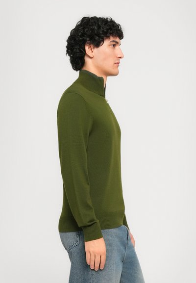 Mann med krøllete mørkt hår iført olivengrønn jakke med glidelås og blå jeans, står i profil mot en enkel hvit bakgrunn.