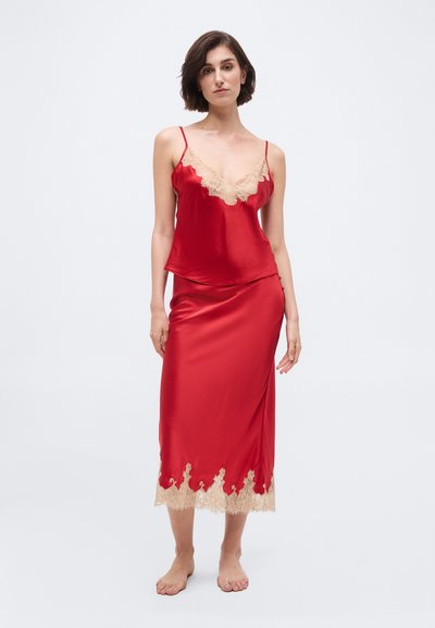 Vrouw staat op blote voeten en draagt een rode satijnen cami met bijpassende midi-rok met beige kanten rand aan halslijn en zoom.