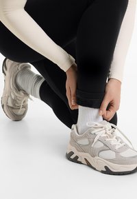 Egy személy igazítja a fekete leggings szárát, hosszú ujjú krémszínű pólót, fehér zoknit és bézs sportcipőt visel egy fehér háttér előtt.