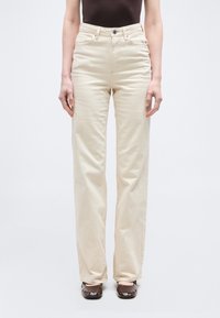 Άτομο που φοράει μπεζ παντελόνι με ψηλή μέση και ίσια γραμμή, με κουμπιά μπροστά, συνδυασμένο με σκούρα καφέ παπούτσια και σκούρο τοπ, στέκεται όρθιο.