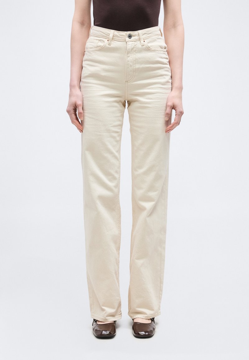 Άτομο που φοράει μπεζ παντελόνι με ψηλή μέση και ίσια γραμμή, με κουμπιά μπροστά, συνδυασμένο με σκούρα καφέ παπούτσια και σκούρο τοπ, στέκεται όρθιο.