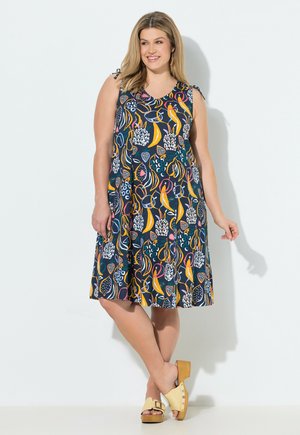 Uśmiechnięta kobieta w kolorowej sukience bez rękawów do kolan z tropikalnym wzorem owoców i beżowych sandałach na koturnie.