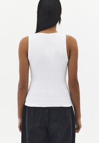Mujer con cabello negro largo que lleva una camiseta sin mangas blanca acanalada y vaqueros negros de talle alto, mostrada de espaldas sobre un fondo blanco.