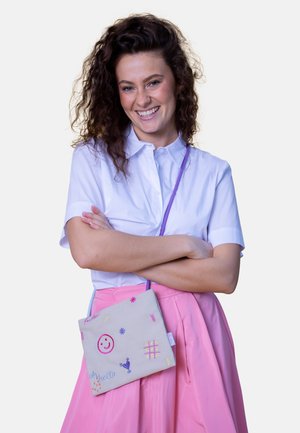 Young woman with curly hair smiling, wearing a white shirt and pink skirt, carrying a small beige crossbody bag with colorful embroidery.
