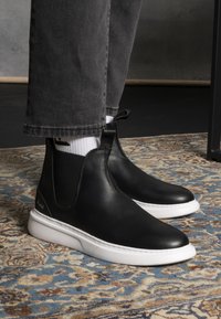 Botas de cuero negras hasta el tobillo con textura suave, paneles laterales elásticos y una suela blanca gruesa. Combinadas con pantalones grises y calcetines blancos.