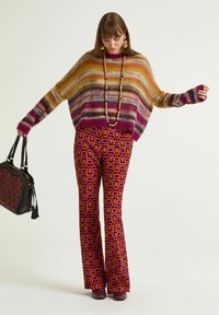 Donna che indossa un maglione a righe peloso, pantaloni a zampa con fantasia, grande collana di perline, orecchini, tiene una borsa nera e marrone, guarda verso il basso.