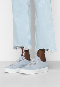 Persona di pelle scura che indossa jeans in denim strappati di un blu chiaro e sneakers in pelle blu chiaro con suola bianca, in piedi su un pavimento grigio.