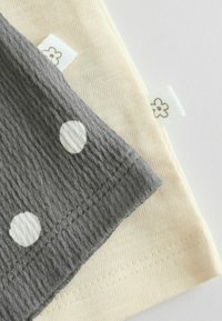 Tissu gris à pois blancs superposé à un tissu tricoté crème, tous deux présentant de petites étiquettes blanches avec des icônes de fleurs simples sur un fond clair.