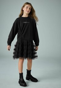 Must mustar ilma kokku pandud külgseamiga, kihiline must tüllseelik, mis sisaldab volangidega kihte, ja paksud mustad pahkluu saapad elastsete külgede jaotustega.