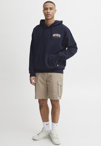 Marineblå hettegenser med kengurulomme og oransje "ARCHIVE" tekst; kombinert med kaki cargo-shorts, hvite sokker og joggesko.