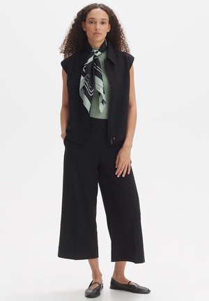 Black sleeveless vest layered over a green textured top, paired with wide black trousers. A patterned black and mint scarf accentuates the outfit.