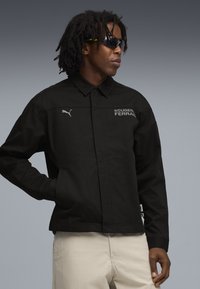 Sort jakke lavet af et glat stof med krave, med teksten "SCUDERIA FERRARI" og et Puma-logo i hvid.