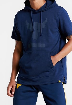 Mann trägt marineblaue kurzärmelige Kapuzenjacke mit erhabenem Stier-Logo und passende marineblaue Hose mit gelbem Stier-Emblem und gelben Seitenstreifen.