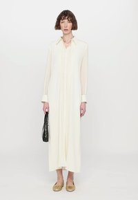 Largo vestido crema con cuello tipo collare, cierre frontal de botones y pliegues verticales. Combinado con alpargatas doradas y un bolso negro.
