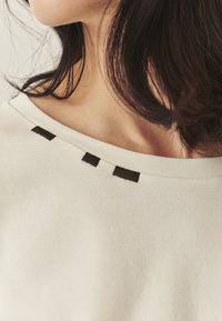 Kremowy top z szerokim dekoltem ozdobiony trzema ciemnobrązowymi akcentami z tkaniny przy kołnierzu. Miękka tekstura i casualowy design.