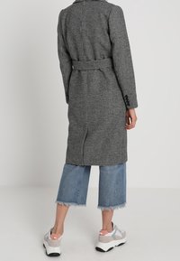 Černobílý kostkovaný dlouhý kabát s opaskem, spárovaný s zkrácenými modrými džíny a robustními teniskami. Hladká textura.