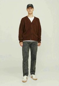 Cardigã castanho tricotado com decote em V e botões, usado sobre uma t-shirt branca. Combinado com jeans de algodão escuro e ténis brancos.