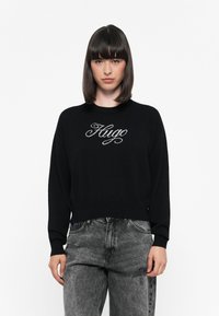 Suéter de punto negro con mangas largas y cuello redondo, que presenta el texto bordado en blanco "Hugo" en el pecho.