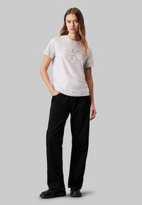 Camiseta de algodón gris claro con detalle de logo en relieve en el pecho, combinada con pantalones de mezclilla negros de cintura alta y zapatos negros.