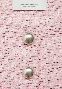 Lyserødt tekstureret stof med vævede flerfarvede tråde; to store sølvknapper placeret foran; plejepose synlig over stoffet.