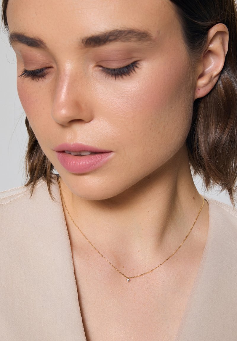 Złoty łańcuszek z małym, przezroczystym zawieszką; modelka ma jasną karnację i nosi beżową bluzkę o subtelnej fakturze.