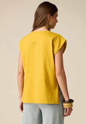 Vrouw met lang bruin haar, gekleed in een mouwloze gele top met kleine tekst op de achterkant en lichtblauwe spijkerbroek, die meerdere armbanden om haar pols draagt.